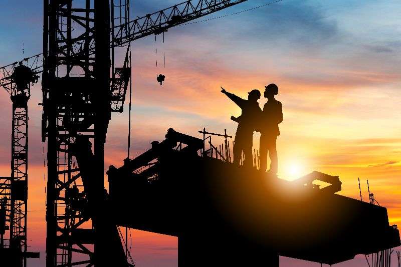 Silhouetted construction workers discussing project plans on a building site at sunset, surrounded by cranes and construction equipment, representing Jacksonville construction services.