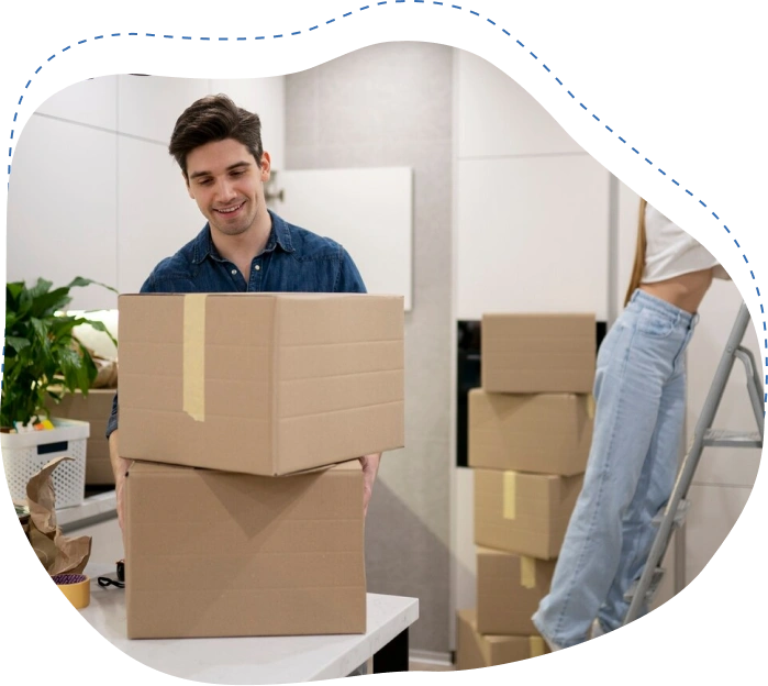 Man smiling while carrying stacked moving boxes in a modern kitchen, preparing for a local move with packing supplies and additional boxes visible in the background.