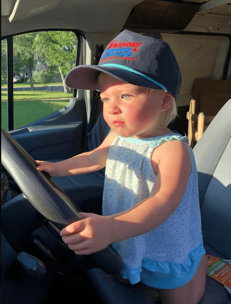 Cute toddler wearing a Skinner Moving & Storage cap sitting at the steering wheel of a moving truck
